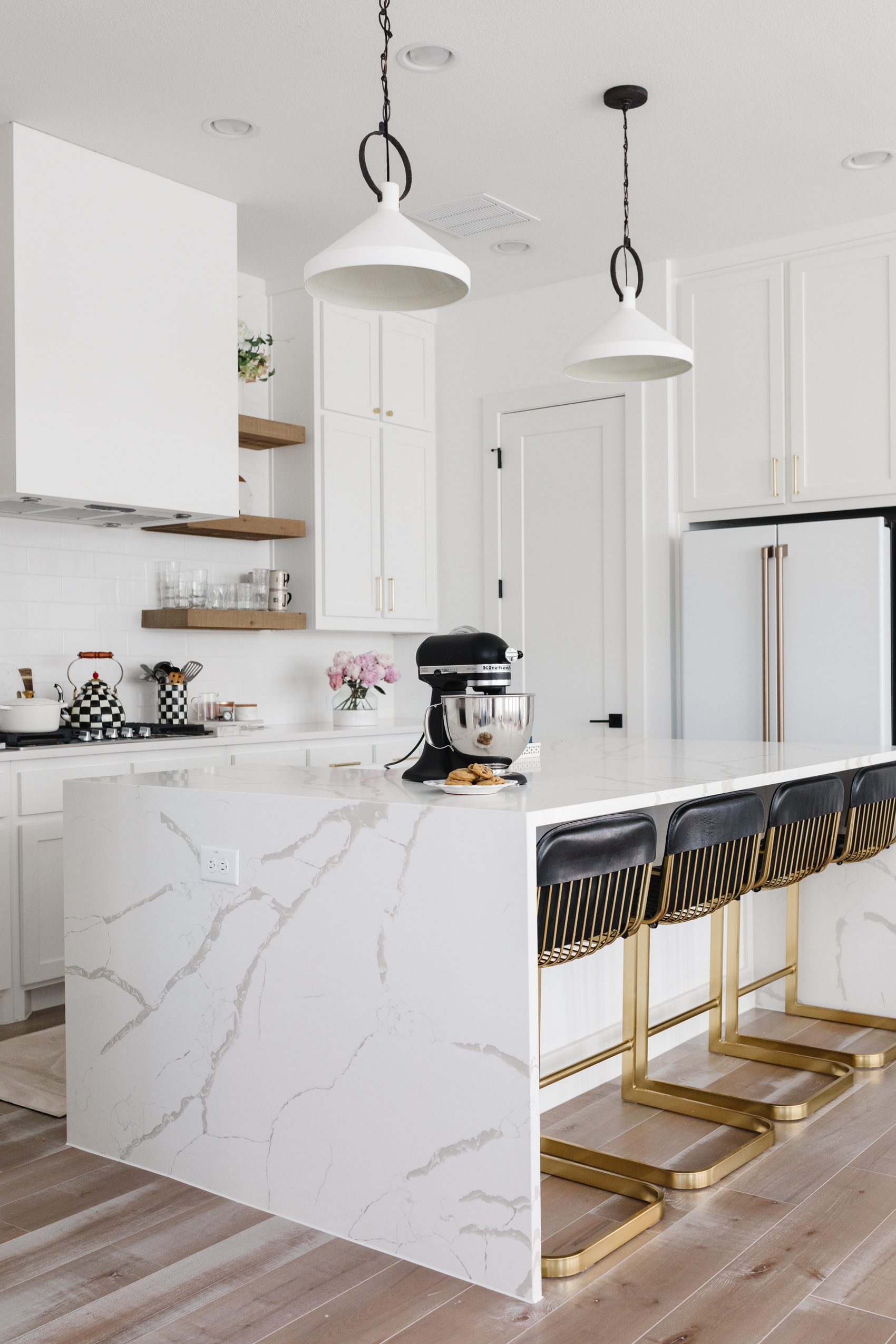 White kitchen with quartz calacatta foresta waterfall island, black CB2 Rake barstools, McGee & Co pendant lights, Cafe Appliances refrigerator, MacKenzie-Childs cookware and more