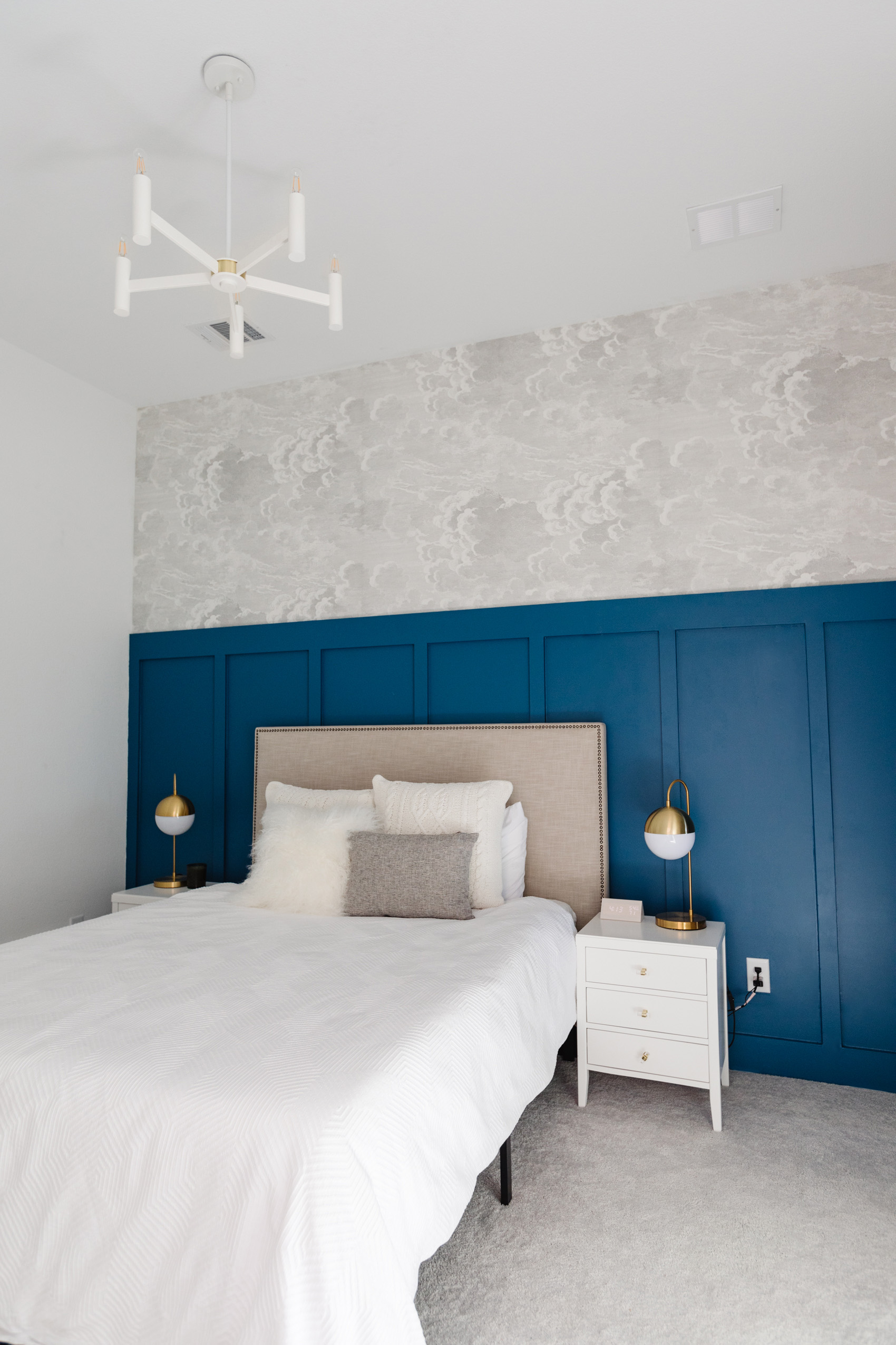 Modern board and batten wall in our guest bedroom with Cole & Son nuvolette wallpaper in a cloud print, white chandelier, upholstered headboard, white nighstands, gold table lamps and a geo duvet