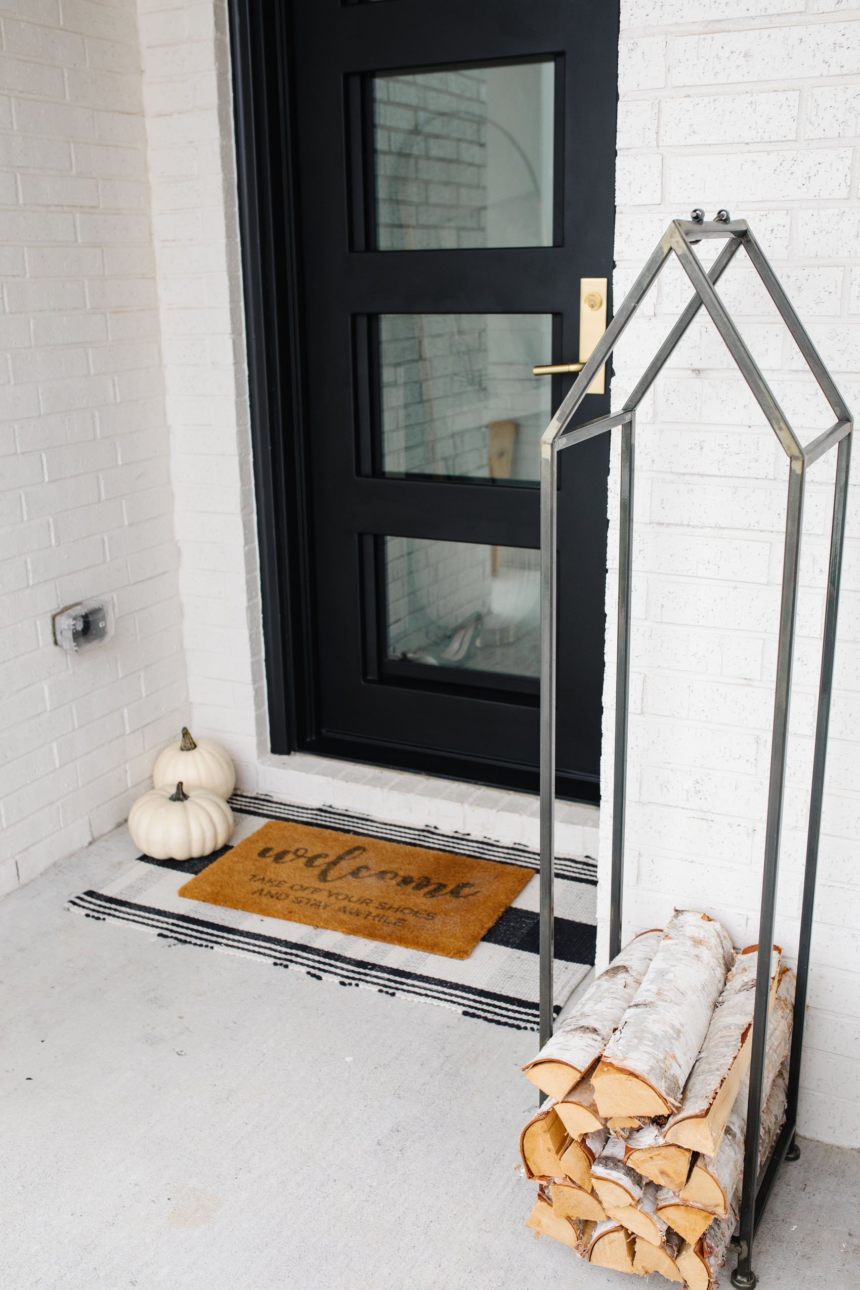 Affordable fall front porch decor with a tartan plaid rug, doormat, white pumpkins, log rack and birch split firewood with an iron front door