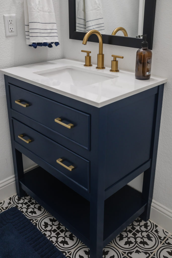 Navy blue small bathroom vanity with a brass faucet and overhead light in a powder bathroom with a black vanity mirror and Serena & Lily hand towels.