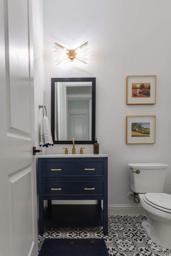 Navy blue small bathroom vanity with a brass faucet and overhead light in a powder bathroom with framed embroidered art and ceramic tile.