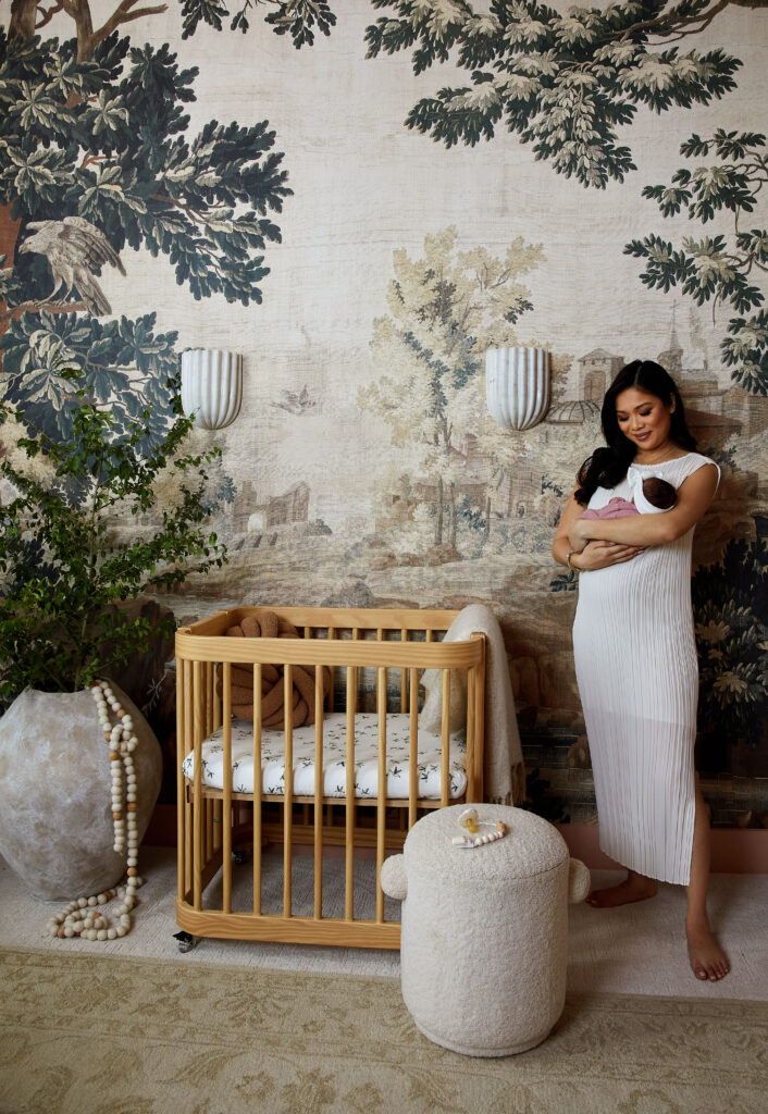 Blogger Hoang-Kim Cung holding her daughter in her modern nursery with mural wallpaper, nestig crib and marble sconces designed by Urbanology Designs