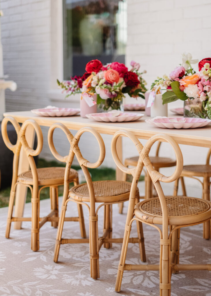 Floral first birthday party theme with a pink backdrop with vibrant flowers, bow wicker kids chairs, kids tables, meri meri scallop paltes and silicone straw cups. Floral rug and fluted pedestal for the cake