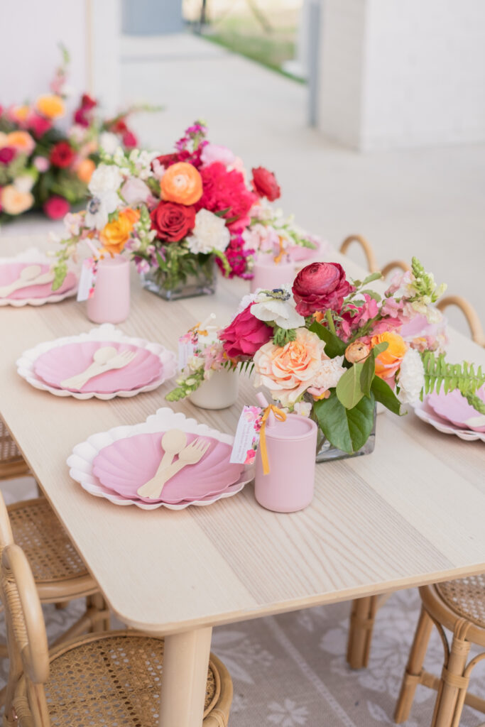 Floral first birthday party theme with a pink backdrop with vibrant flowers, bow wicker kids chairs, kids tables, meri meri scallop paltes and silicone straw cups. Floral rug and fluted pedestal for the cake